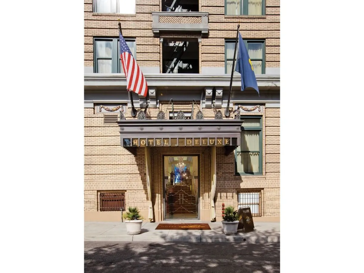 An elegant exterior shot featuring the entrance of Hotel Deluxe, complete with flag displays and decorative architectural details. The image captures the inviting and classic facade, emphasizing the hotel's connection to Portland's cultural heartbeat.