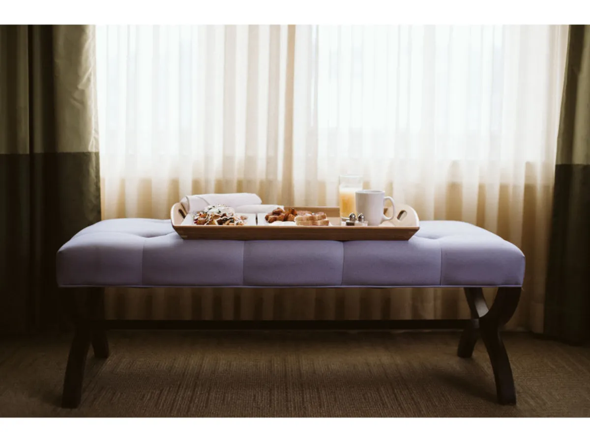 A cozy breakfast-in-bed setup featuring a tray with breakfast items atop a plush bed, accompanied by a purple upholstered bench and bright curtains. The atmosphere is tranquil, ideal for restful mornings and quiet downtime.