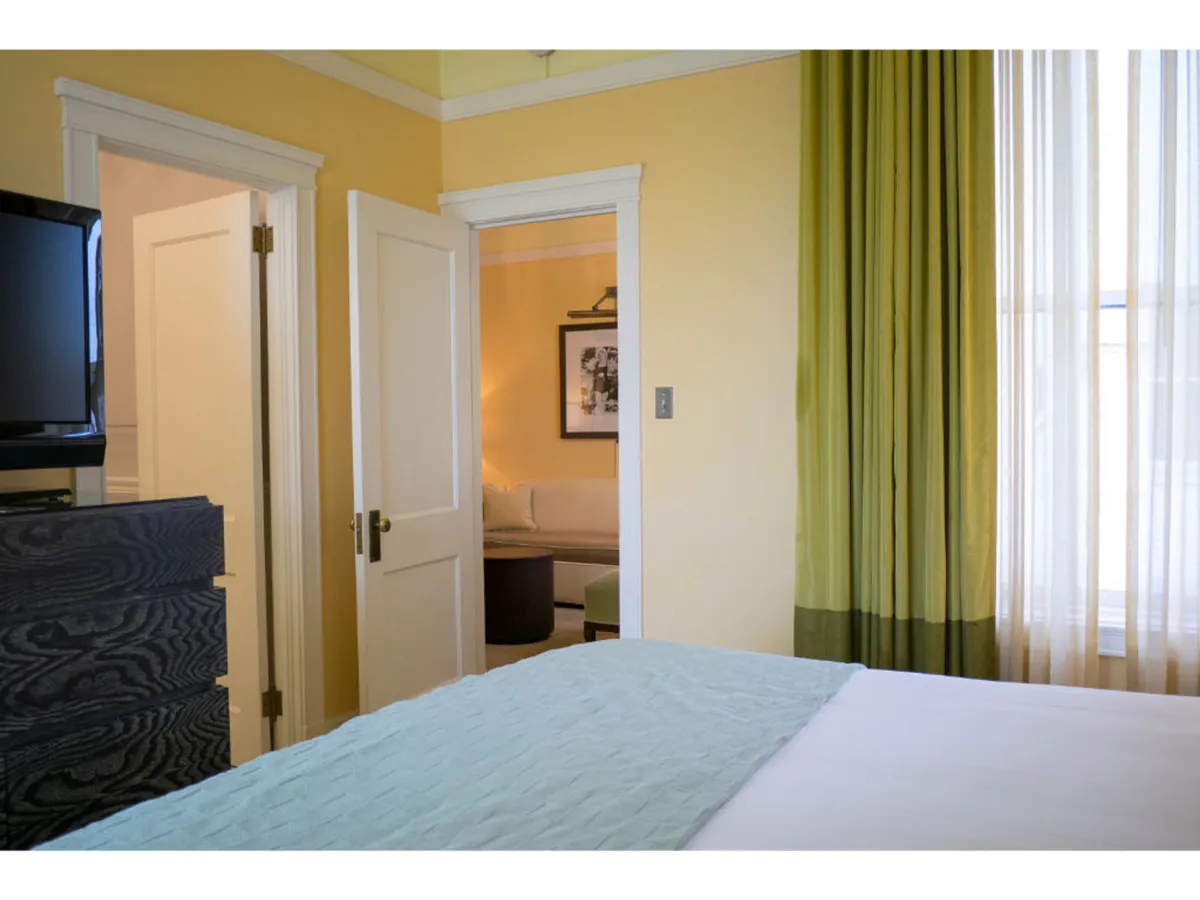 A spacious, inviting bedroom highlighted by a bed with a light blue cover, green curtains, a modern TV unit, and a visible doorway leading to an adjoining space. The room blends comfort and functionality, suggesting space for both relaxation and privacy.
