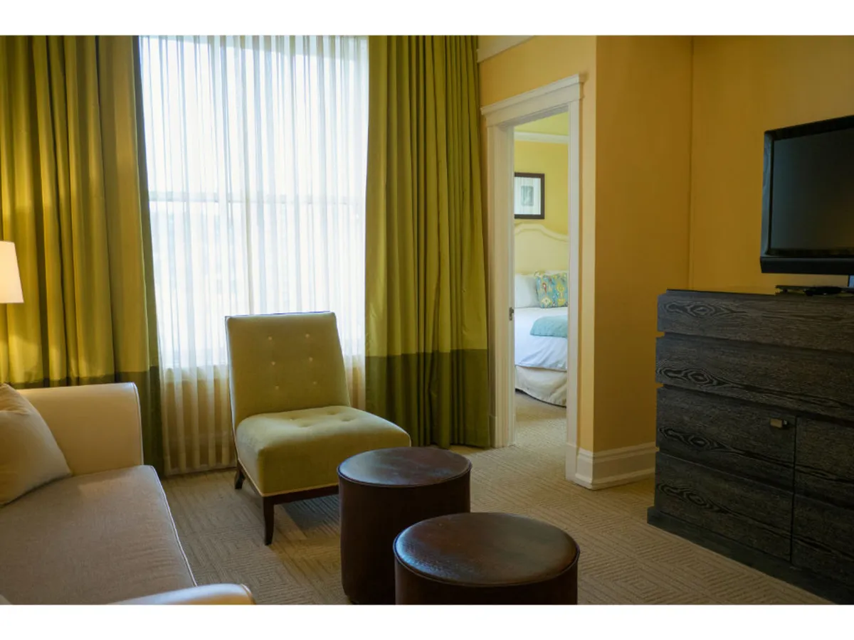 An inviting corner of the living area with a large window and sheer curtains, a green armchair, wooden side tables, and a TV unit. The setting is ideal for reading, watching TV, or relaxing in natural light, evoking comfort and versatility for both leisure and work.