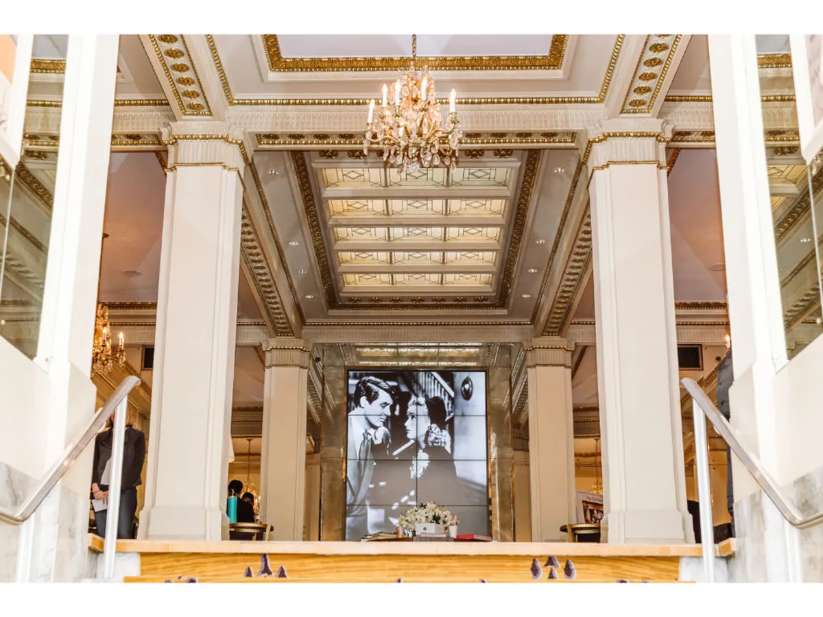 A refined interior scene featuring a grand chandelier, ornate ceiling, and a sweeping staircase. The space is anchored by a large screen displaying a historical image, suggesting a venue ready for formal gatherings, ceremonies, or galas. The overall atmosphere is celebratory and sophisticated, ideal for memorable events.