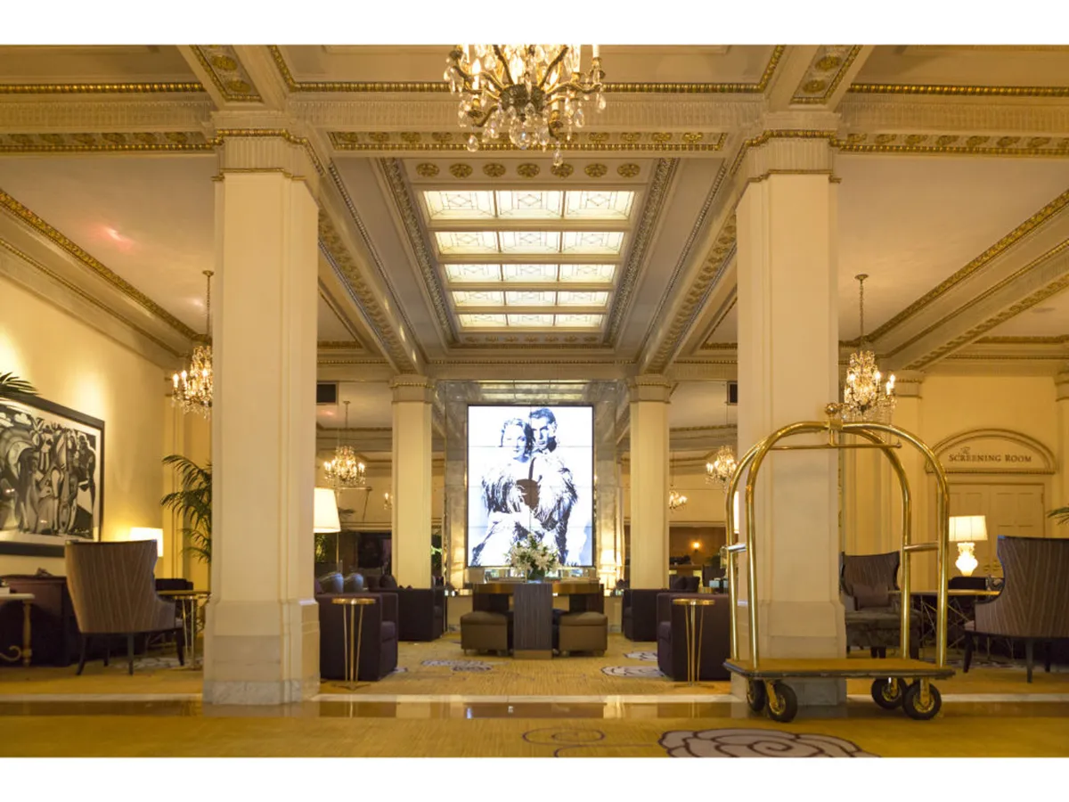 A chic interior lounge with sparkling chandeliers, comfortable seating, curated artwork, and a central screen. The setting is lively, with guests socializing and appreciating local art, embodying the blend of sophistication and creativity the hotel offers.