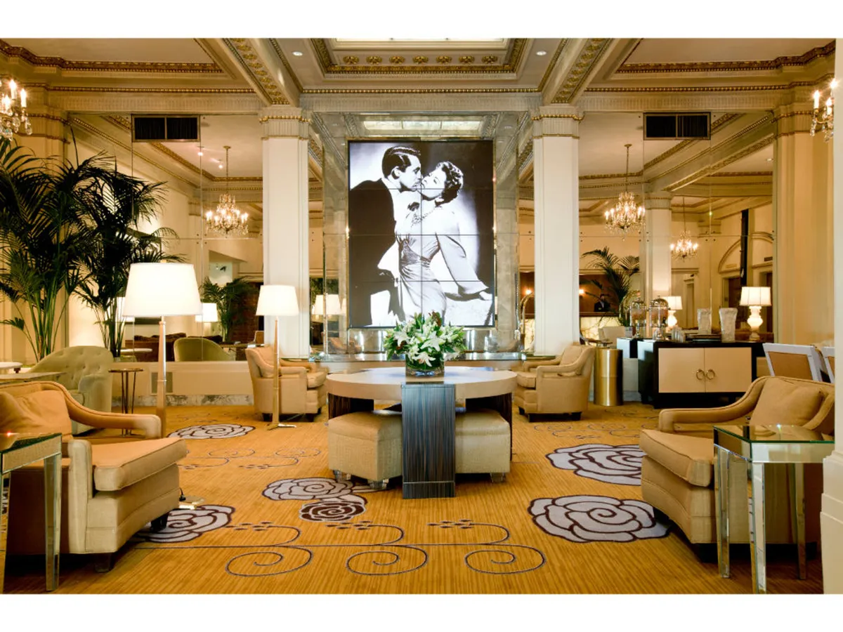 A sophisticated seating area features a large black and white artwork of a couple, a central round table with a floral arrangement, and plush, stylish furnishings. Guests converse, enjoy drinks, and relax, highlighting the versatile, welcoming nature of the space.
