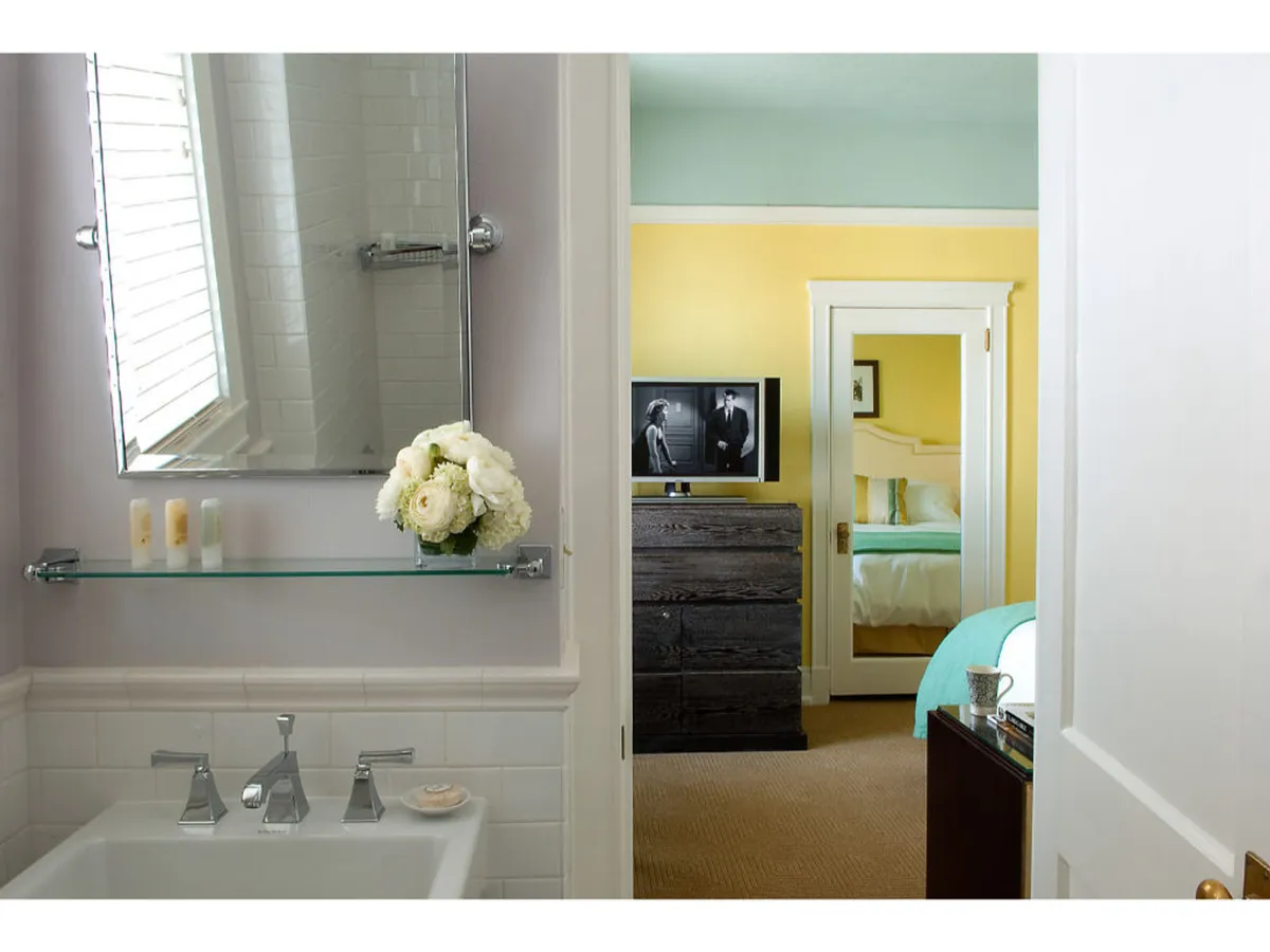 This image features a tranquil bathroom setting highlighted by a large mirror with a floral arrangement, a visible dresser, and a television set seen from the bedroom. The scene evokes a sense of calm and relaxation, with soft lighting and thoughtful decor that suggest a luxurious, spa-like experience. The mirror, a key focal point, hints at the availability of amenities like a beauty/shaving mirror.