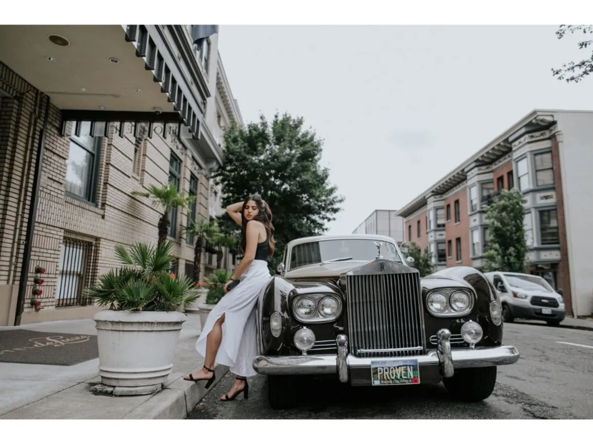 The image features a vintage car parked near the hotel entrance, a woman dressed in white, and stylish planters. The urban setting and architectural backdrop suggest a lively scene that blends elegance with the excitement of city exploration.