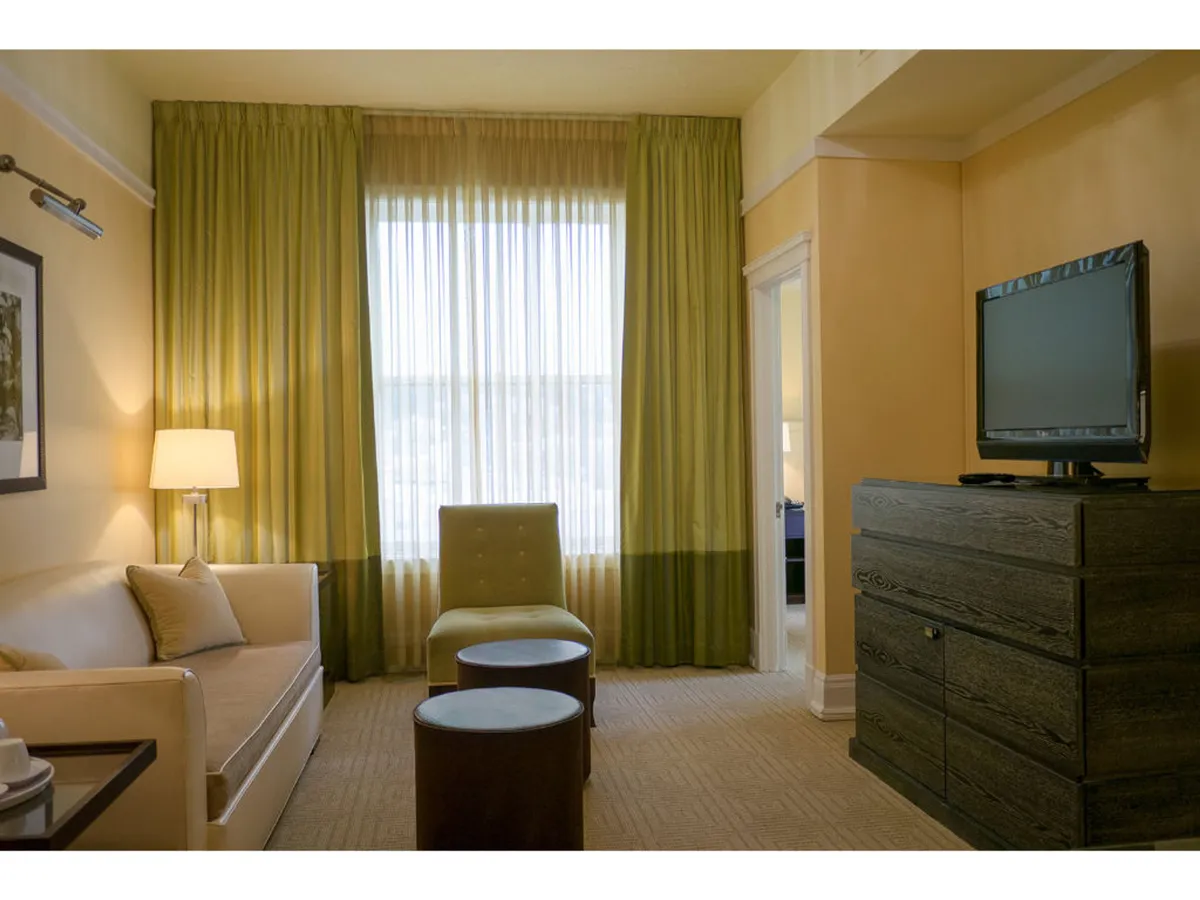 A spacious living area filled with natural light from large windows with sheer curtains. The room is appointed with a plush sofa, decorative armchairs, a lamp, and a 38-inch HD TV, creating an inviting setting for relaxation or entertainment. Subtle vintage accents and elegant furnishings contribute to an Old Hollywood-inspired ambiance.