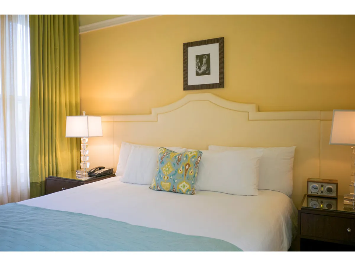A serene bedroom scene centered on a king-sized bed adorned with decorative pillows, flanked by elegant lamps and rich drapes. A framed picture hangs above the headboard, and natural light filters through the window, emphasizing plush linens and inviting textures.