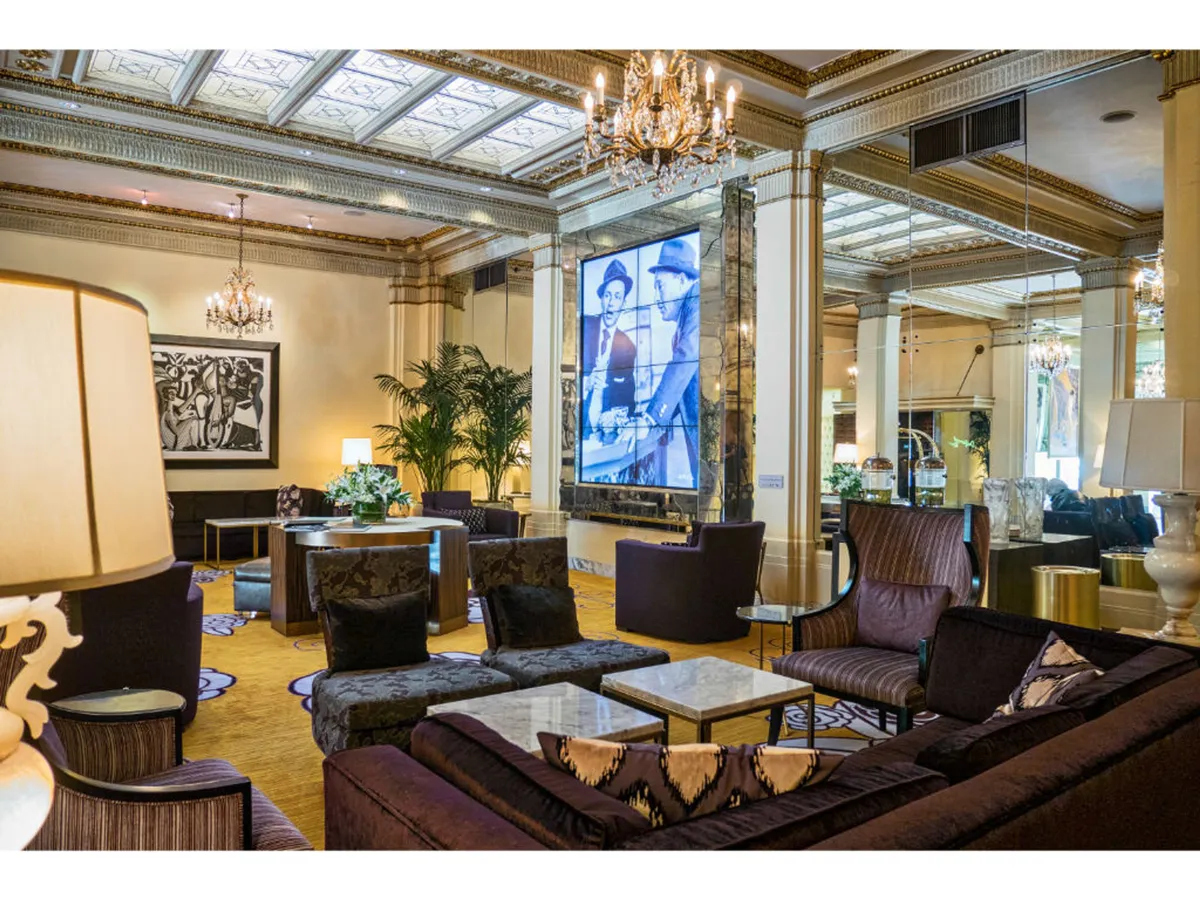A chic and spacious interior lounge featuring a glowing chandelier, a modern art piece, and a large screen displaying a historical image. The area is furnished with plush seating arranged for both intimacy and group gatherings, inviting guests to relax, socialize, or appreciate the artful ambiance.