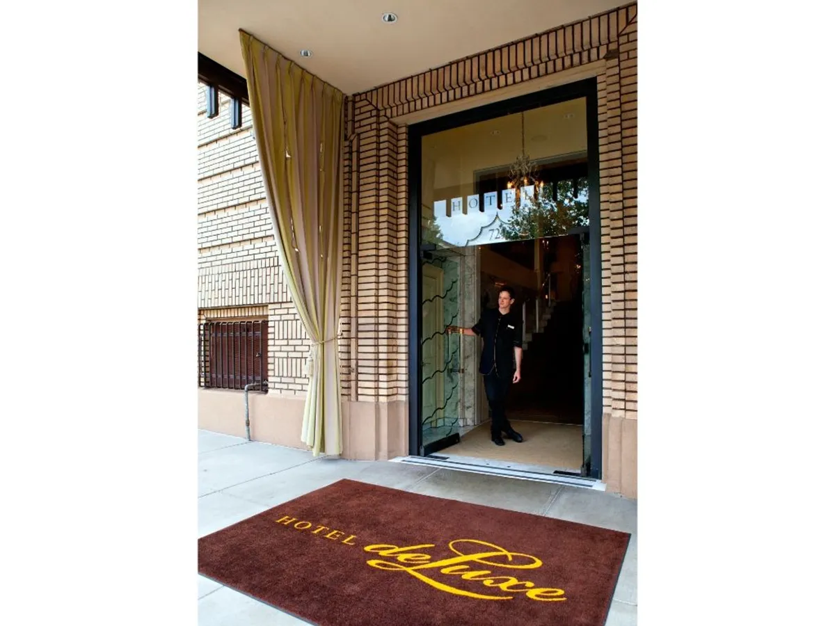 The image shows the entrance door to Hotel deLuxe, adorned with a doormat bearing the hotel's name. A staff member is visible, welcoming guests at the threshold, creating a sense of hospitality and anticipation for what's inside. The setting suggests the beginning of an experience, with tasteful architectural details framing the doorway.