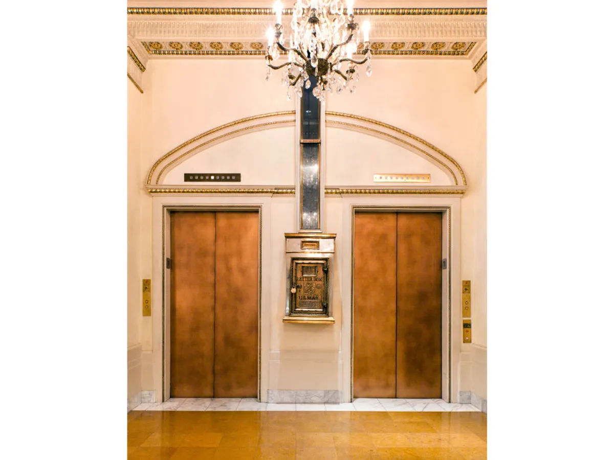 A beautifully appointed lobby space with elegant elevators, a dazzling chandelier, and a decorative archway. The mood is both luxurious and nostalgic, capturing guests as they move through the heart of the hotel, ready to explore or return to their suite.