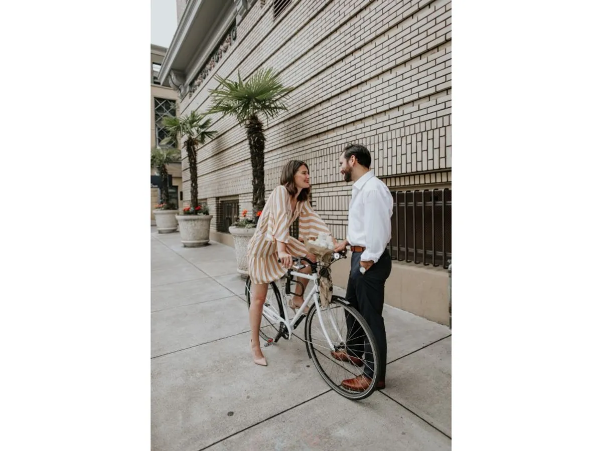 An exterior scene with a couple smiling at each other, a bicycle adorned with flowers, and lush potted plants. The image suggests active, pet-friendly exploration and a touch of boutique charm, tying in the complimentary bike rental and the hotel’s welcoming atmosphere.